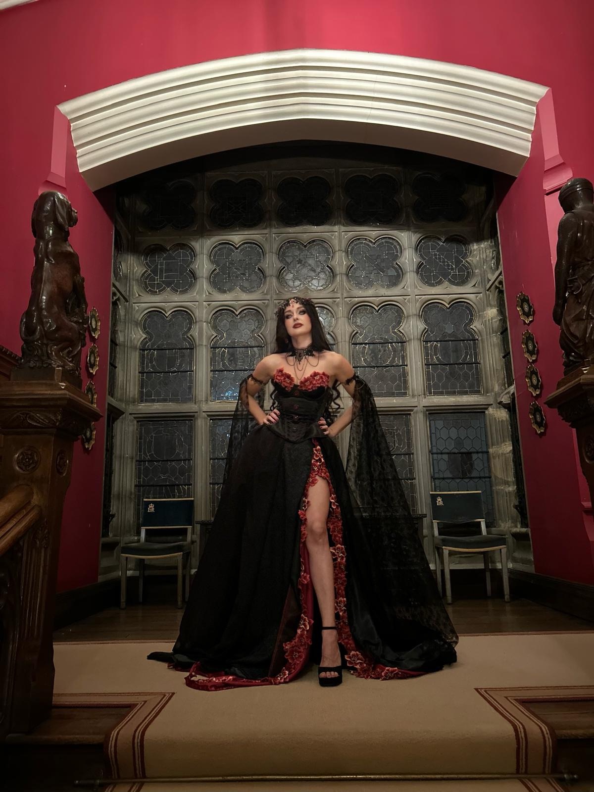 The finished Vampire Queen gown: a black corset with red roses, a full black skirt with a front slit lined in red, sheer black lace sleeves and a train spreading across the floor.
