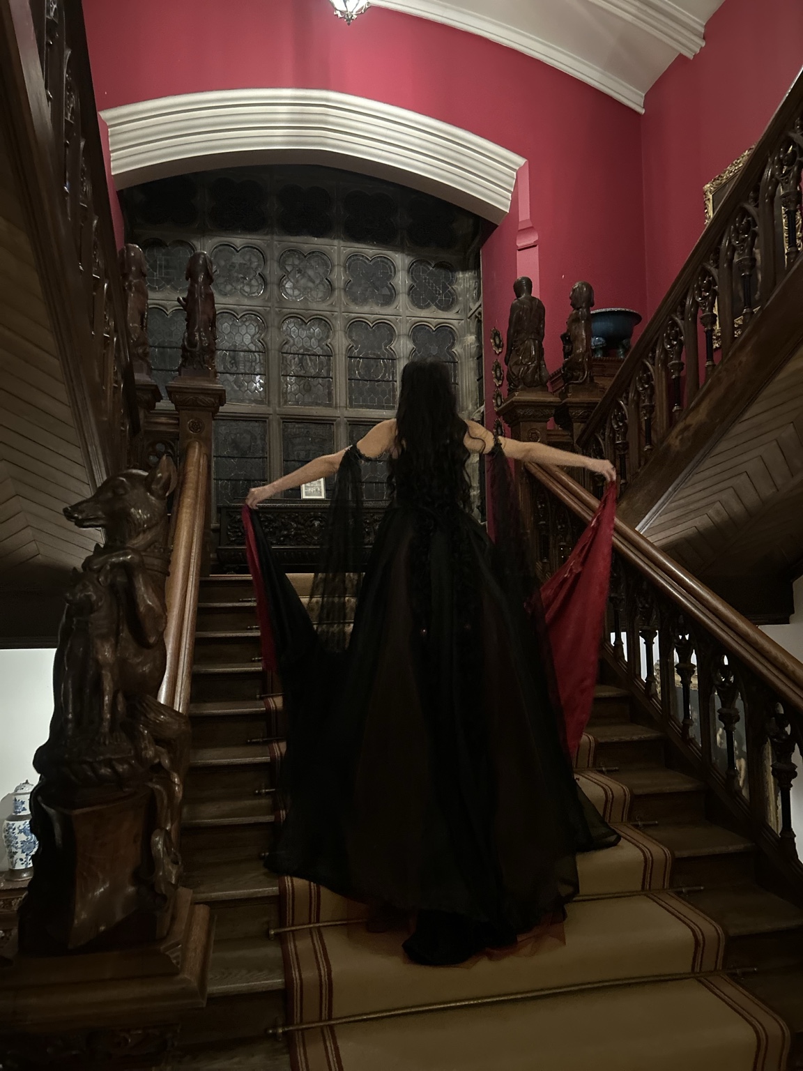 Seen from behind on a carved wooden staircase, with the black and red train and lace sleeves spread out.